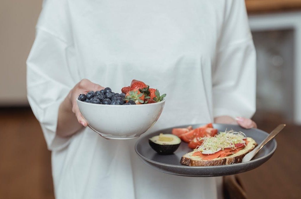 Person holding a bowl of fresh berries and a plate with avocado toast, displaying a healthy breakfast spread.