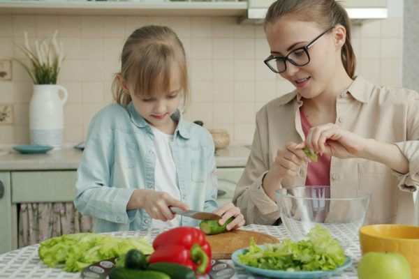 Adorable little girl is helping mother in kitchen cutting vegetables for salad and talking making dinner together. Healthy nutrition, lifestyle and people concept.
